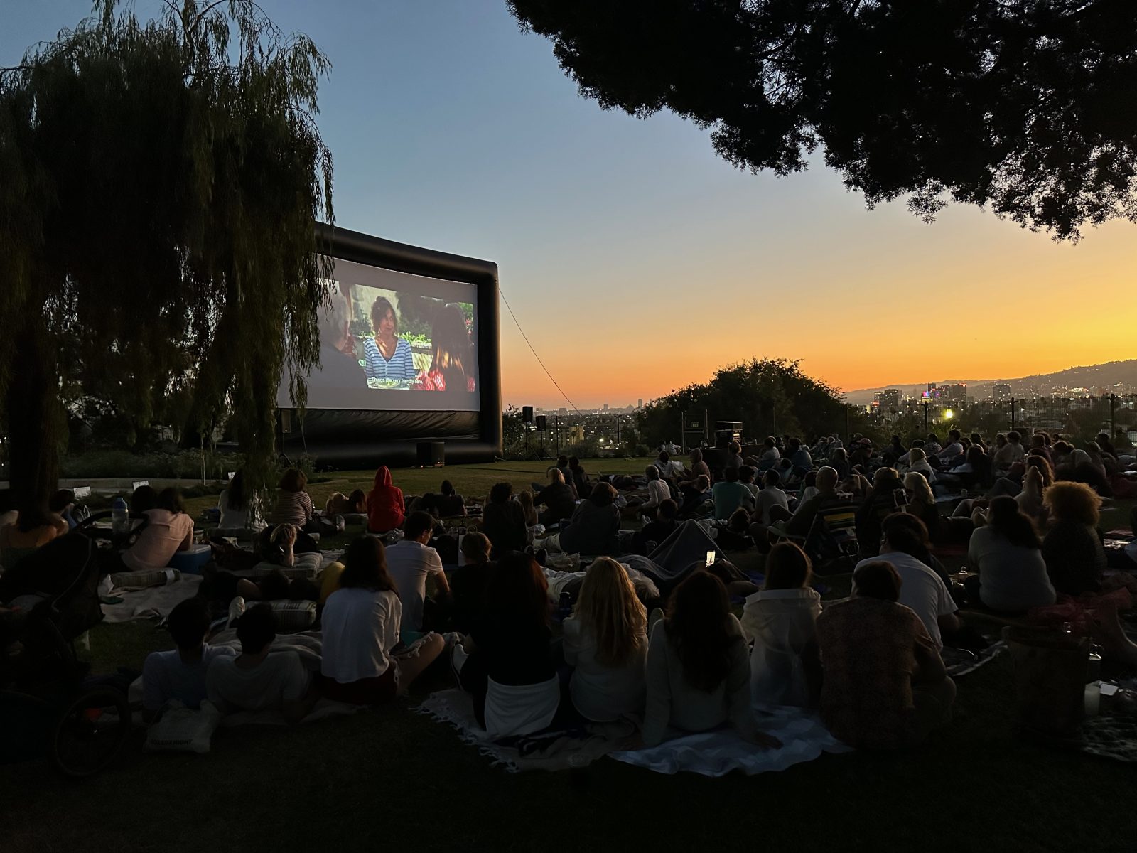 Crowd sitting on outdoor lawn watching a movie with sunset in the background.